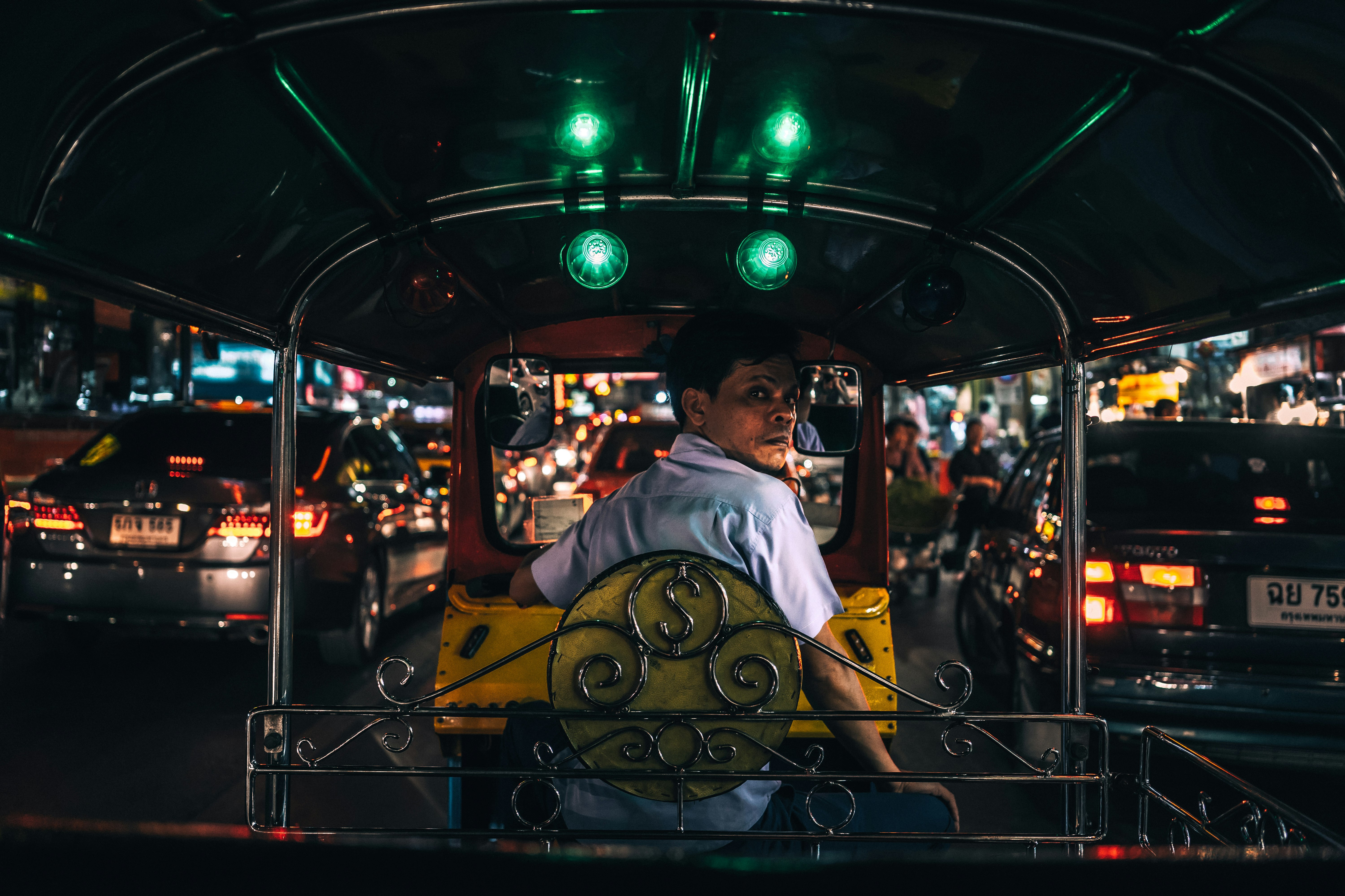 man in white collared shirt sitting in yellow and black autorickshaw between cars with taillights turned on at night